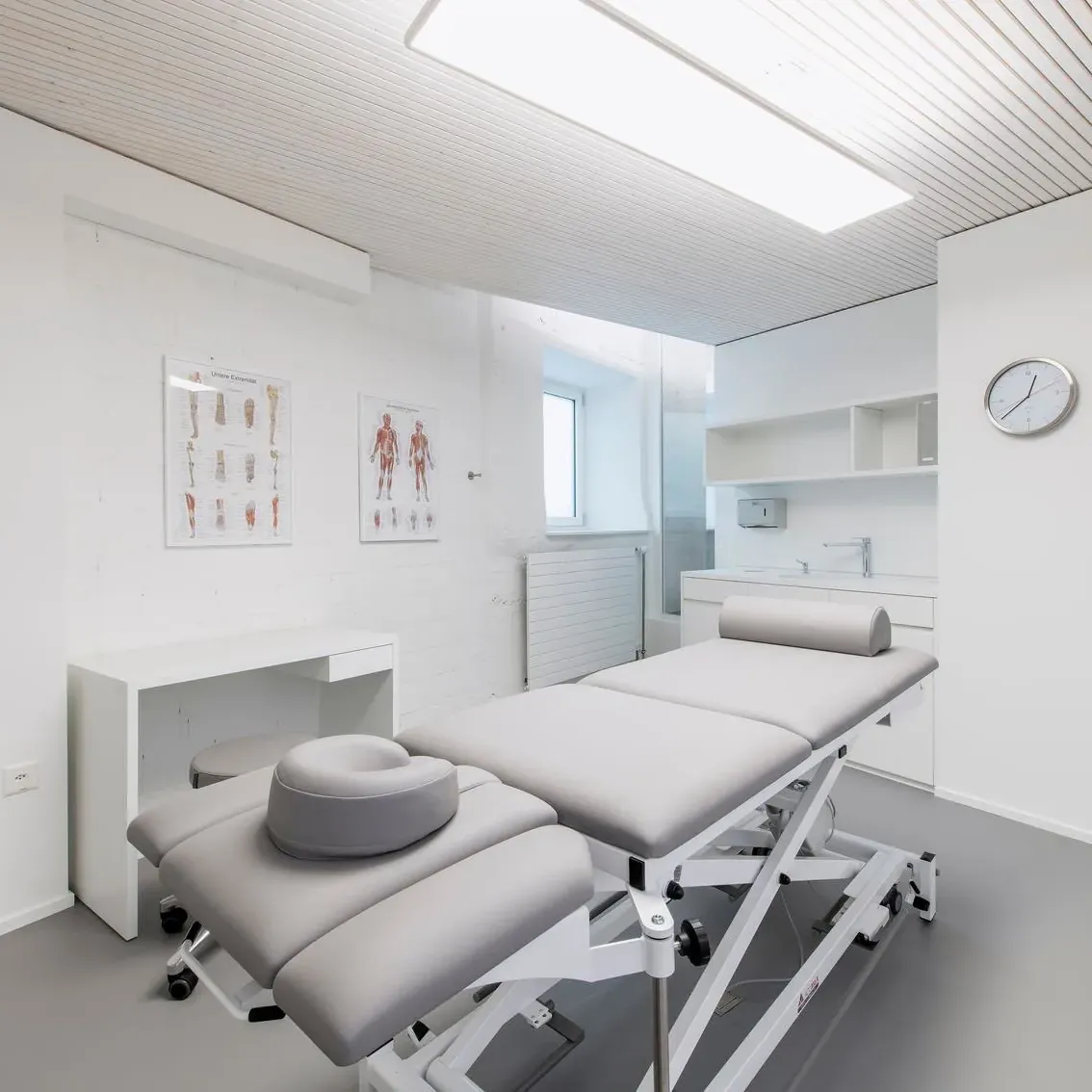 Private Healthcare treatment room with bed in centre of the room, clock on the wall and ribbed acoustic ceiling in off white